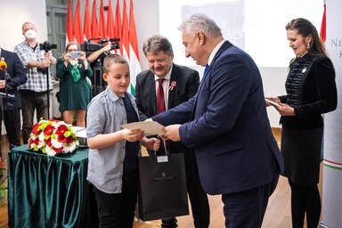 Budapest, 2022. m�rcius 17.
H�lka Arnold (b) �tveszi az elismer�s�t Semj�n Zsolt minisztereln�k-helyettest�l (j) �s  Sz�sz Jen�t�l, a Nemzetstrat�giai Kutat�int�zet eln�k�t�l a Magyar z�szl� �s c�mer rajz- �s essz�p�ly�zat d�j�tad� �nneps�g�n Budapesten,
